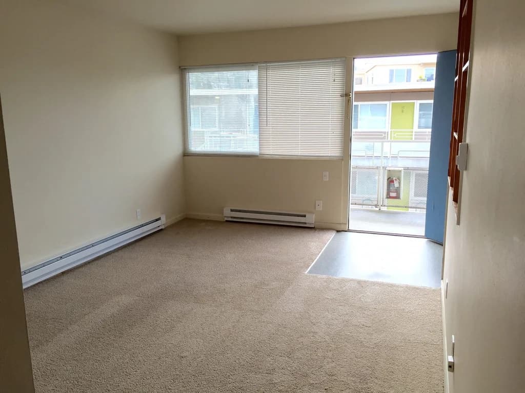 Empty living room with light carpet, a window with blinds, and door leading to a balcony.