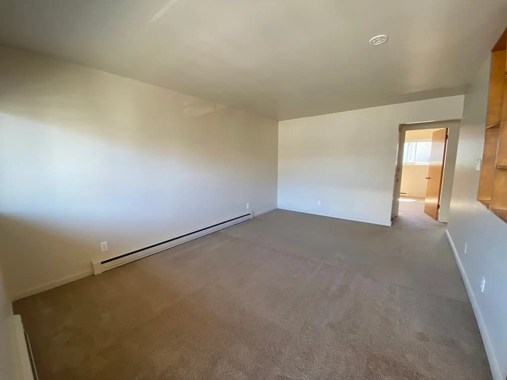 Empty living room with beige carpet, light walls, and baseboard heating, with a doorway to anothe...