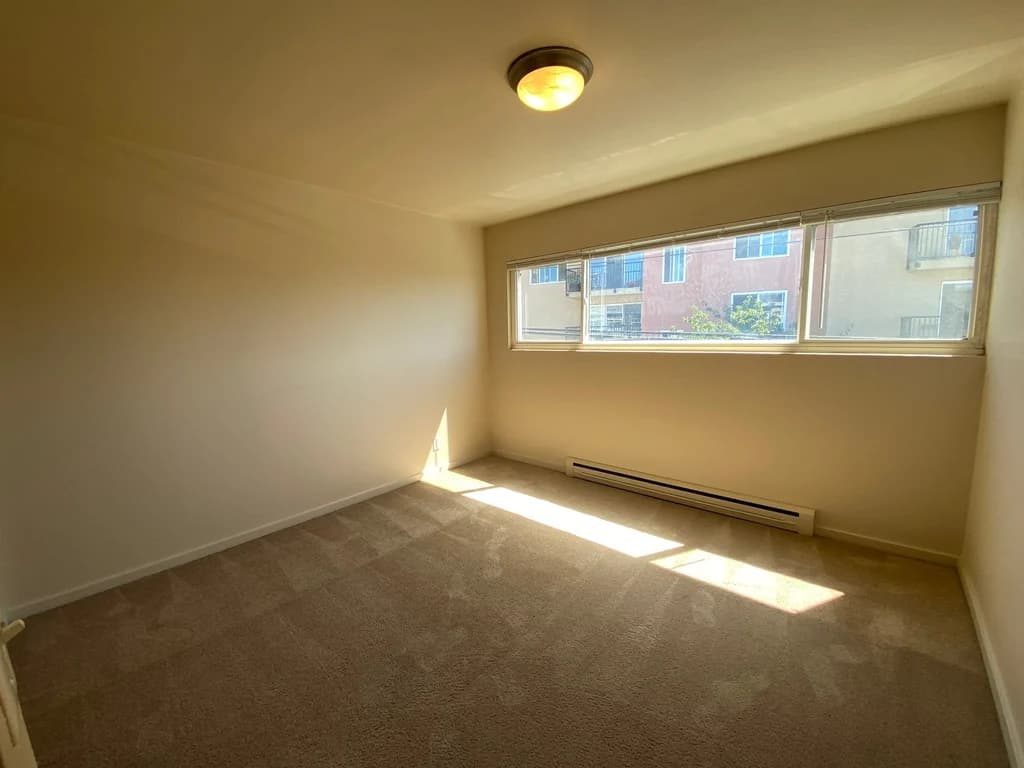 Empty carpeted bedroom with large window, blinds, and natural light streaming onto the floor.