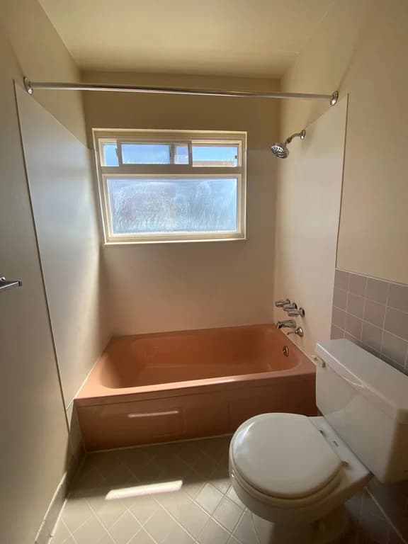 Bathroom with a peach bathtub, shower, window, and white toilet at Cortina Apartments.