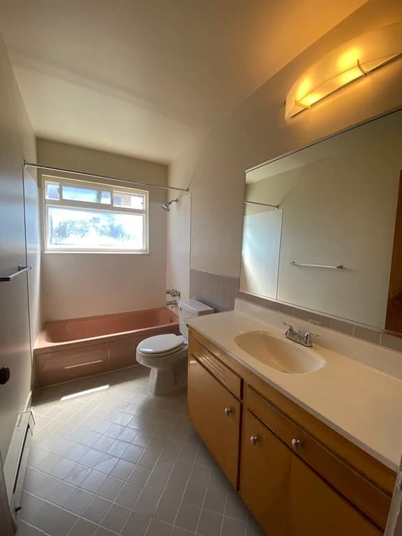 Bathroom with a light pink bathtub, toilet, wood vanity, white sink, large mirror, and window.