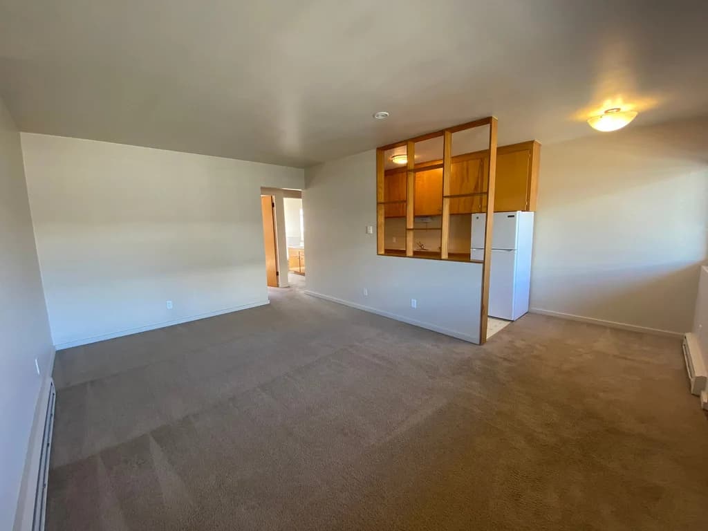Carpeted living room at Cortina Apartments, with a partial wall looking into a kitchen area.
