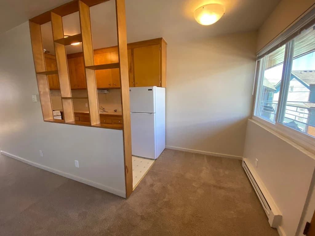 Open living area with carpet, large window, wooden room divider, and view into kitchen with fridg...